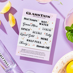 A collection of dissolvable drink markers from Glass Tats laid out on a table with a glass of water and lemon slices. The markers have various playful phrases written on them. Available at Be Charmed