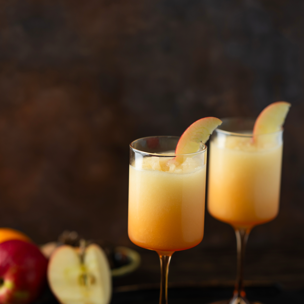 Two glasses of orange cocktails with apple slices on a dark background. Available at Be Charmed