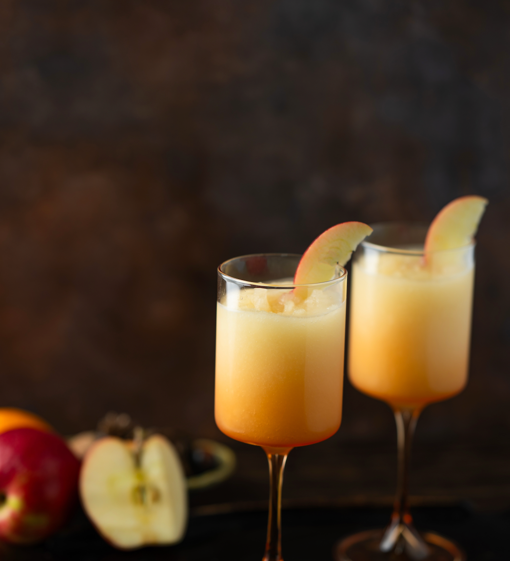 Two glasses of orange cocktails with apple slices on a dark background. Available at Be Charmed
