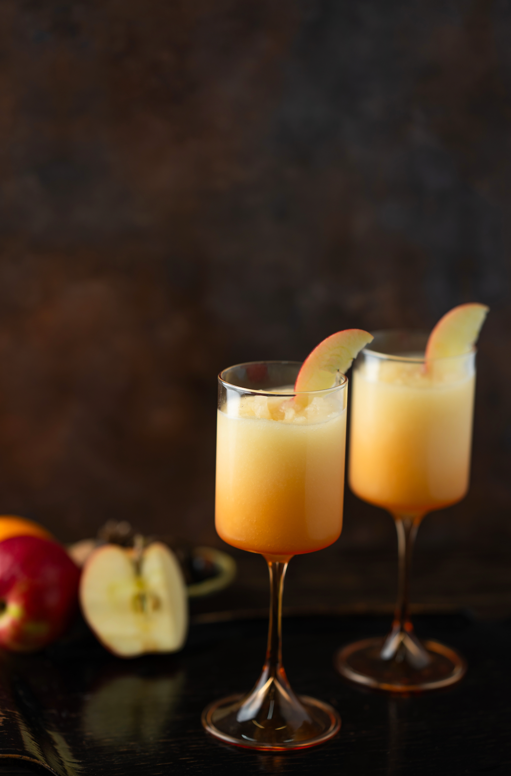 Two glasses of orange cocktails with apple slices on a dark background. Available at Be Charmed