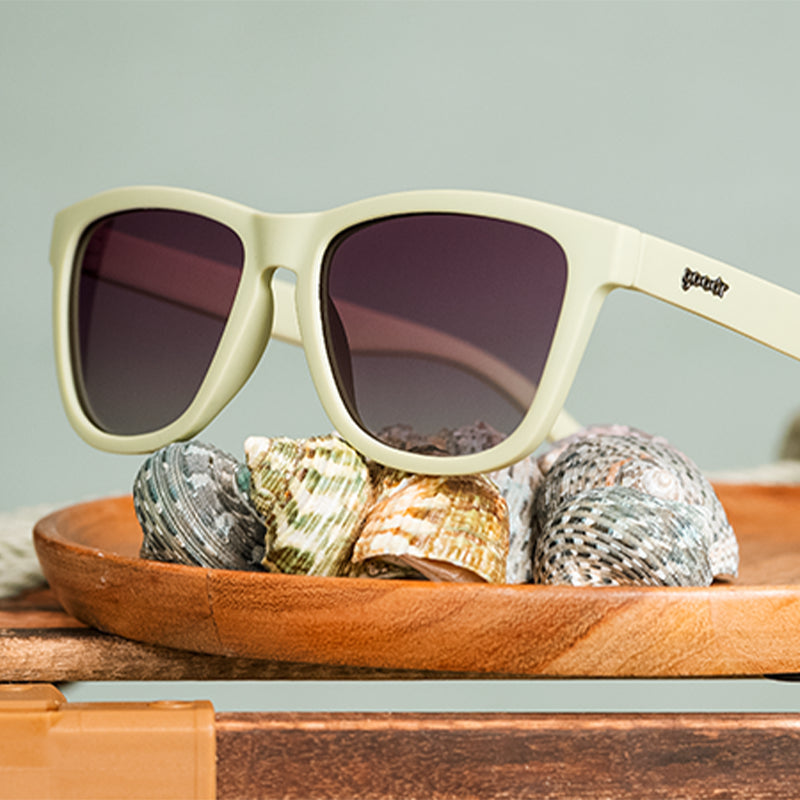 A pair of sage green Goodr sunglasses with purple gradient lenses placed on a wooden surface, with textiles and other accessories around. Available at Be Charmed