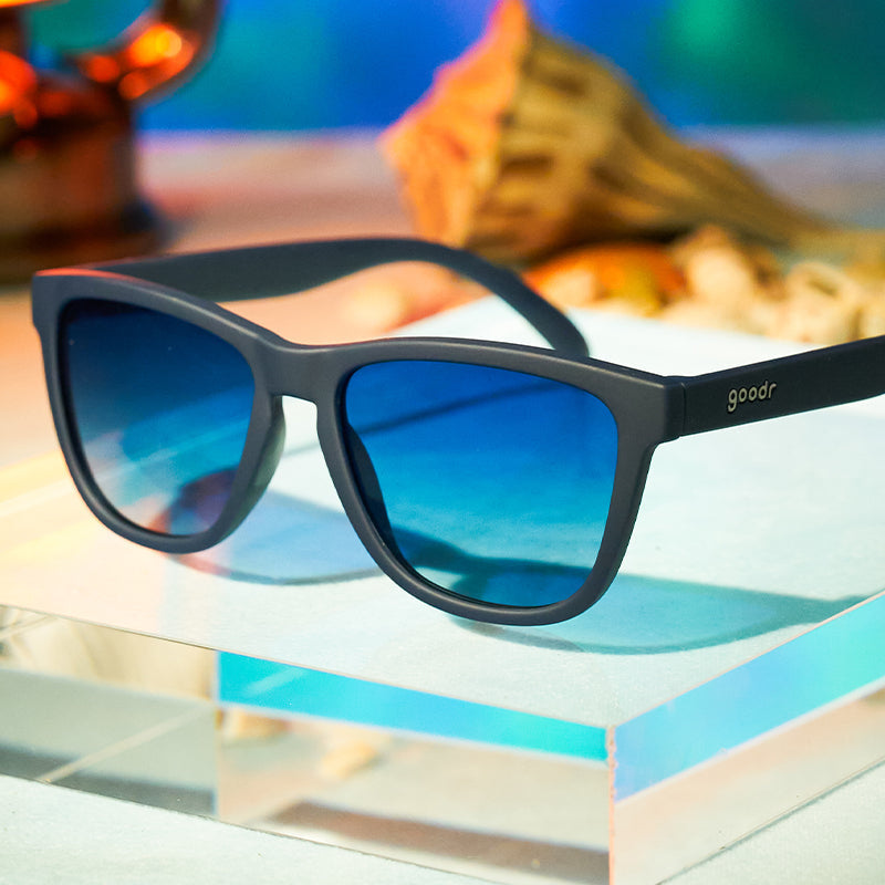 Goodr Drinks Seawater, Sees Future sunglasses sitting on book, featuring polarized reflective lenses and ocean-inspired frames—performance and style at Be Charmed.
