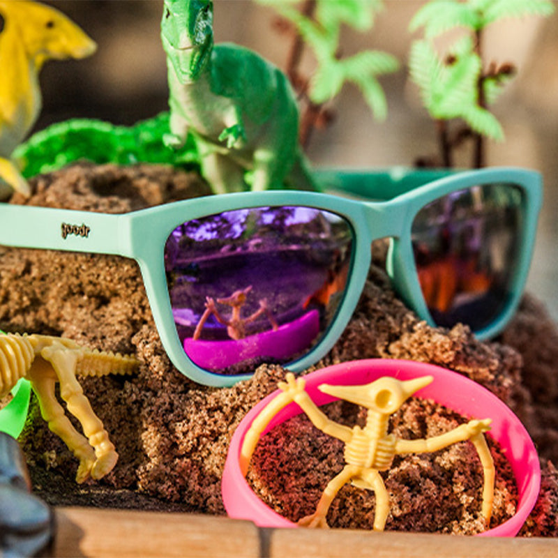 Goodr Electric Dinotopia Carnival Sunglasses sitting on beach sand with plastic dinosaurs. Available at Be Charmed