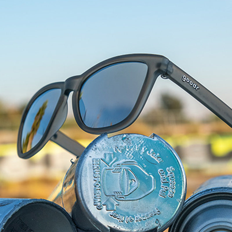 Goodr Going To Valhalla Witness Sunglasses Propped on bottles featuring polarized black lenses and a no-slip frame, inspired by Viking grit and perfect for outdoor adventures, Available at Be Charmed