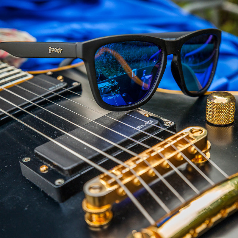 Goodr Mick & Keith's Midnight Ramble Sunglasses on guitar strings featuring classic black frames and blue smoke-tinted polarized lenses, designed for high-energy adventures and effortless cool, available at Be Charmed