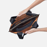 Brown and black HOBO leather handbag held by a person on a light gray background. Available at Be Charmed