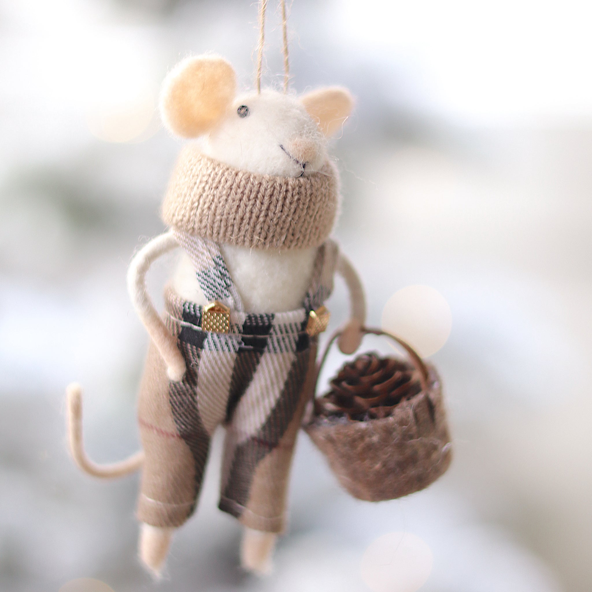 Decorative mouse ornament wearing a sweater and plaid skirt, holding a basket, against a blurred natural background. Available at Be Charmed