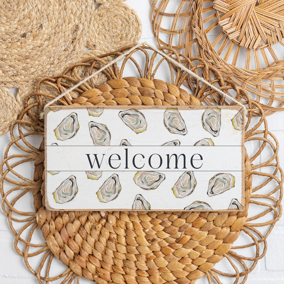 Decorative sign with 'welcome' text and oyster design on a woven mat. Available at Be Charmed