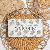 Decorative sign with 'welcome' text and oyster design on a woven mat. Available at Be Charmed