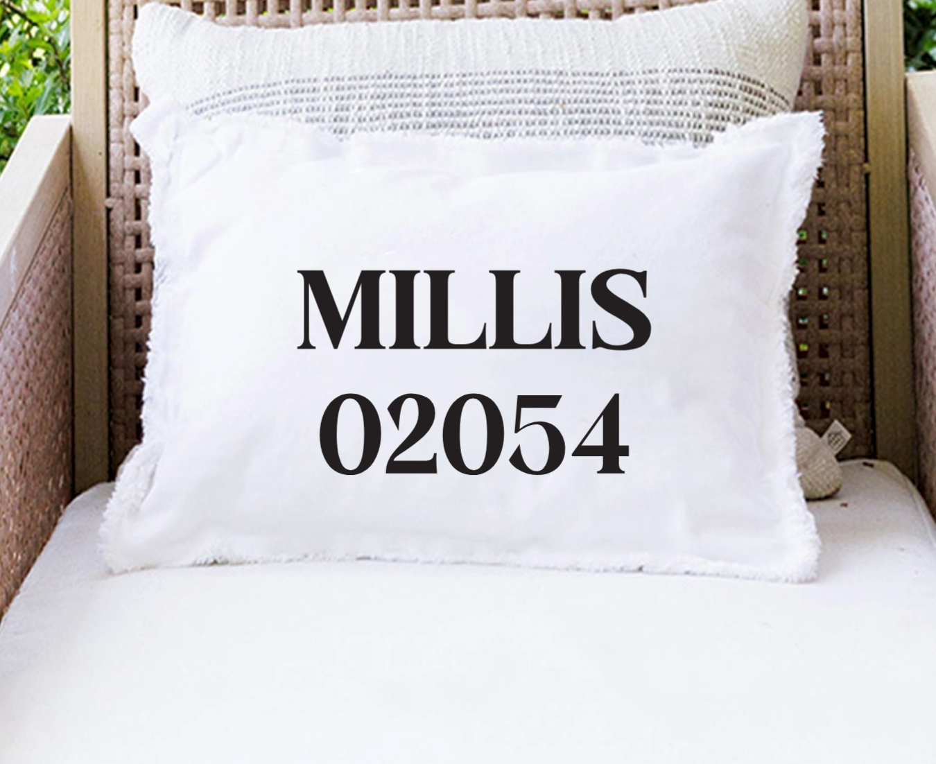 A white throw pillow with fringed edges printed with the black text 'MILLIS 02054' in a bold serif font, resting on a white cushion in front of a woven rattan chair. Available at Be Charmed