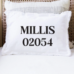 A white throw pillow with fringed edges printed with the black text 'MILLIS 02054' in a bold serif font, resting on a white cushion in front of a woven rattan chair. Available at Be Charmed