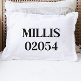 A white throw pillow with fringed edges printed with the black text 'MILLIS 02054' in a bold serif font, resting on a white cushion in front of a woven rattan chair. Available at Be Charmed