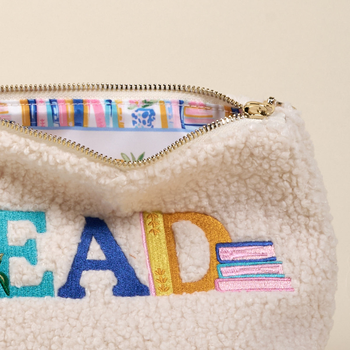 Cream-colored fleece pouch with colorful 'READ' text and striped interior. Available at Be Charmed