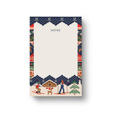 A vertical Rifle Paper notepad with a winter ski-themed border design. The top section is cream-colored with the word 'NOTES' printed in gray, while the bottom and side borders feature a colorful pattern of skiers, a cabin, a tree, and snow in navy, red, green, and beige tones. Available at Be Charmed
