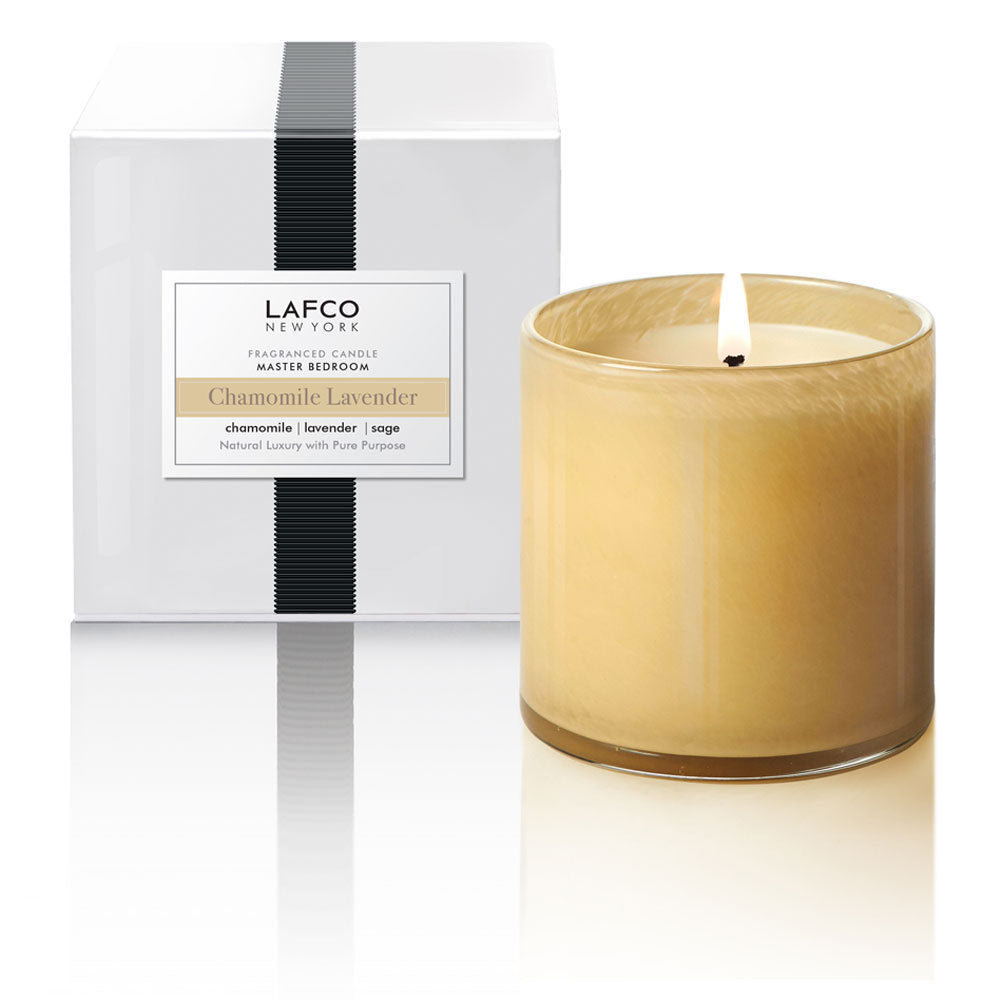 A beige candle with a label reading 'LAFCO - Chamomile Lavender' in a clear handblown art glass vessel, accompanied by a white box with a lid. Available at Be Charmed