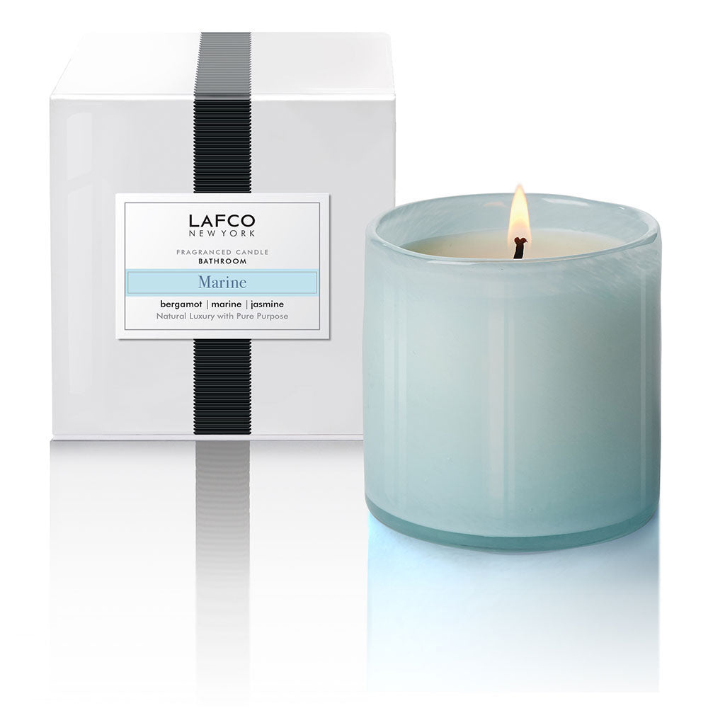 A LAFCO brand marine-colored candle in a clear art glass vessel, with a label indicating the scent of marine and jasmine. The candle is placed in front of a white box with the LAFCO logo. Available at Be Charmed
