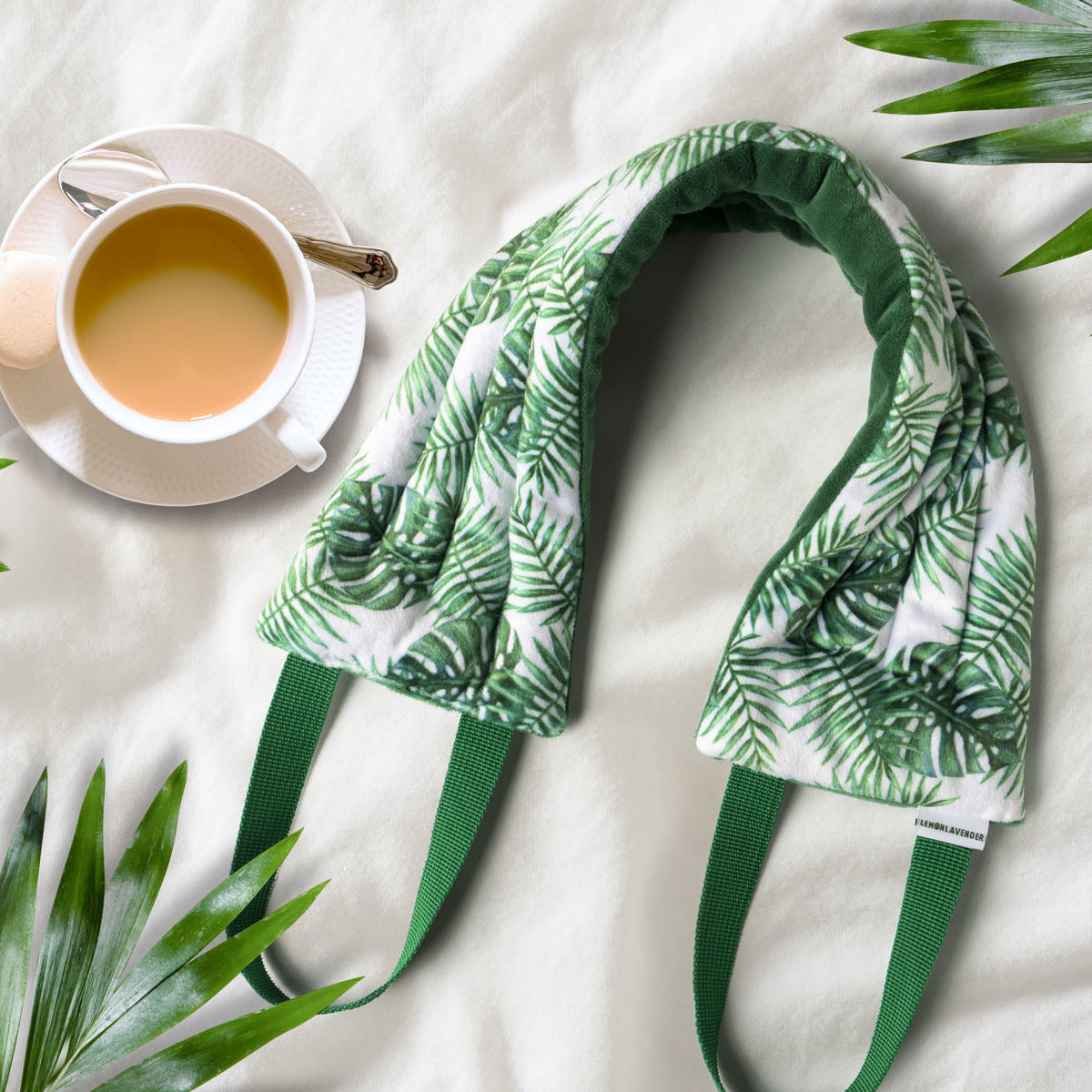 A micfiber neck wrap with a lemon and lavender design, displayed on a flat surface with a cup of tea and green leaves in the background.. Available at Be Charmed
