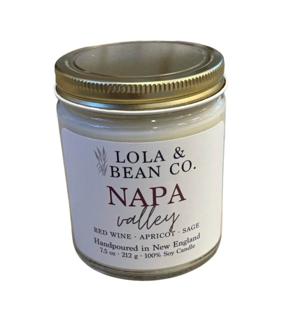 A white candle jar with a gold lid labeled 'Lola & Bean Co., Napa Valley' with scents listed as red wine, apricot, sage. Available at Be Charmed