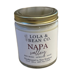 A white candle jar with a gold lid labeled 'Lola & Bean Co., Napa Valley' with scents listed as red wine, apricot, sage. Available at Be Charmed