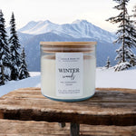A 3-wick soy candle with a bamboo lid, labeled 'LOLA & BEAN CO. WINTER WOODS', placed on a wooden surface with a snowy mountain background. Available at Be Charmed