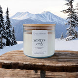 A 3-wick soy candle with a bamboo lid, labeled 'LOLA & BEAN CO. WINTER WOODS', placed on a wooden surface with a snowy mountain background. Available at Be Charmed