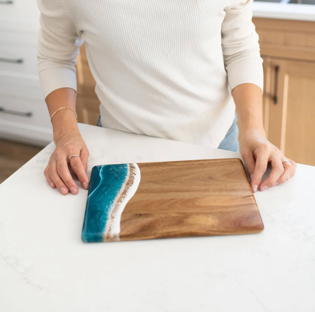 Person holding a wooden cutting board with a blue resin design on a kitchen counter. Available at Be Charmed