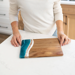 Person holding a wooden cutting board with a blue resin design on a kitchen counter. Available at Be Charmed