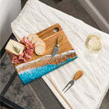 Lynn & Liana's small cheese board with a wood and resin design, featuring ocean colors at the bottom. It is accompanied by an assortment of meats, cheeses, and a glass of white wine, presented on a white cloth. Available at Be Charmed
