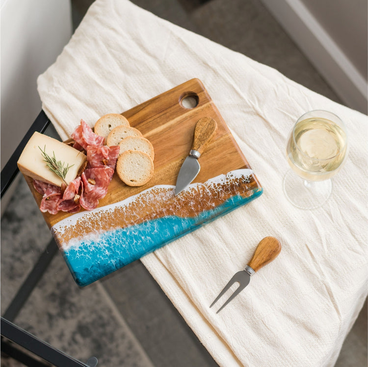 Lynn & Liana's small cheese board with a wood and resin design, featuring ocean colors at the bottom. It is accompanied by an assortment of meats, cheeses, and a glass of white wine, presented on a white cloth. Available at Be Charmed