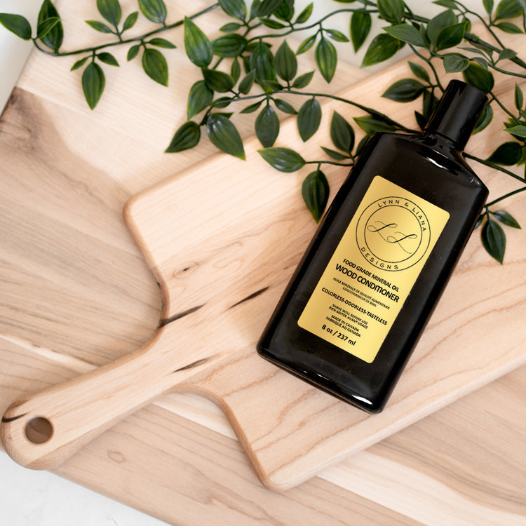 A bottle of Lynn & Liana Wood Board Oil placed on a wooden surface with a wooden spatula and green leaves in the background. Available at Be Charmed