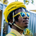 A man wearing yellow and blue Goodr sunglasses with blue reflective lenses and a black helmet with the logo 'velotezze' on it. Available at Be Charmed