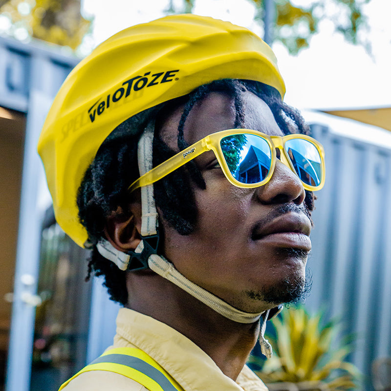 A man wearing yellow and blue Goodr sunglasses with blue reflective lenses and a black helmet with the logo 'velotezze' on it. Available at Be Charmed