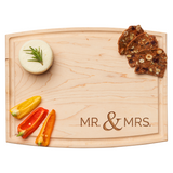 Wooden cutting board with 'MR. & MRS.' engraving, accompanied by vegetables and crackers. Available at Be Charmed