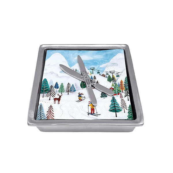 A silver-colored Mariposa napkin box with a ski design, featuring crossed skis and a snowy mountain backdrop. Available at Be Charmed