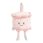 A plush toy resembling a happy cake with a pink velvet body, white frosting, and multicolored sprinkles. It has a striped candle on top and is standing on two legs. Available at Be Charmed