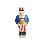 A ceramic figurine of Uncle Sam in a blue coat and hat with a red bow tie, standing on a round base which is not visible in the image. Available at Be Charmed