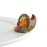 A small, decorative cornucopia-shaped dish charm, featuring autumn-colored fruits such as pumpkin and cranberry, placed on a white plate. Available at Be Charmed