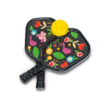 A mini pickleball paddle with a colorful floral and fruit design on a black background, featuring a yellow pickleball in the center. Available at Be Charmed
