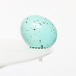 A blue speckled egg-shaped mini charm on a white stand, resembling a Easter egg. Available at Be Charmed