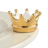 A gold-colored crown-shaped charm placed on a white plate. Available at Be Charmed