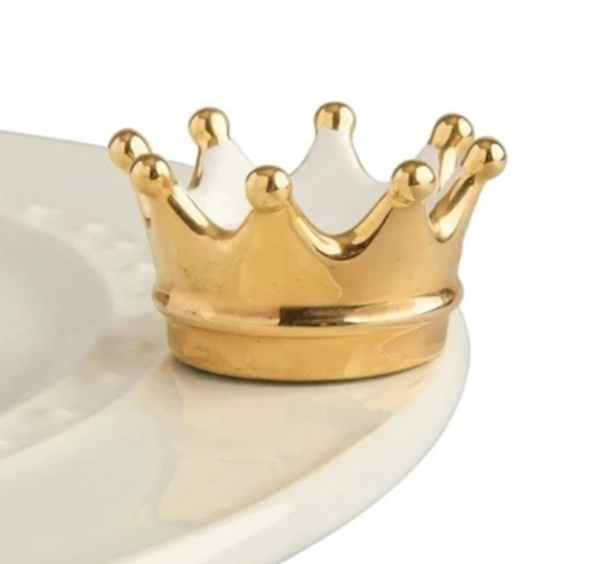 A gold-colored crown-shaped charm placed on a white plate. Available at Be Charmed