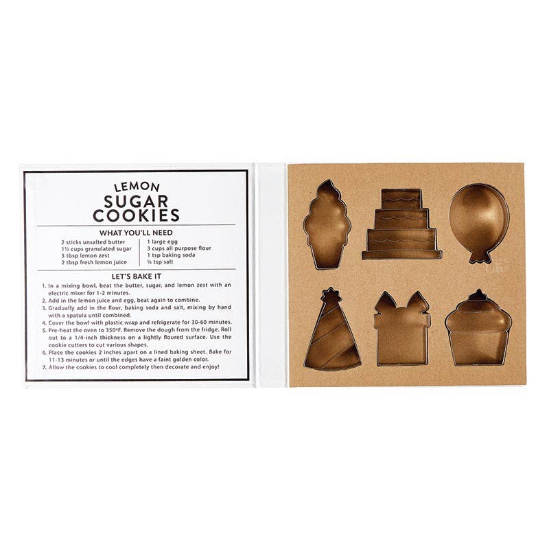 Open box of Creative Brands Birthday cookie cutter set in a magnetic book-style box with 6 stainless steel shapes (cake, balloon, candle, present, party hat, heart) and a lemon sugar cookie recipe inside lid—Available at Be Charmed