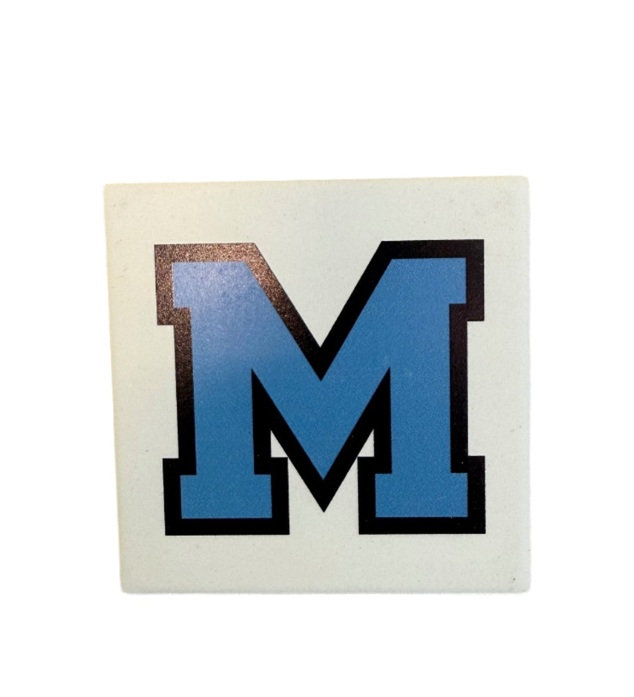 A square ceramic coaster with a cork base, predominantly white with a large blue 'M' logo in the center and black outline detailing. Available at Be Charmed