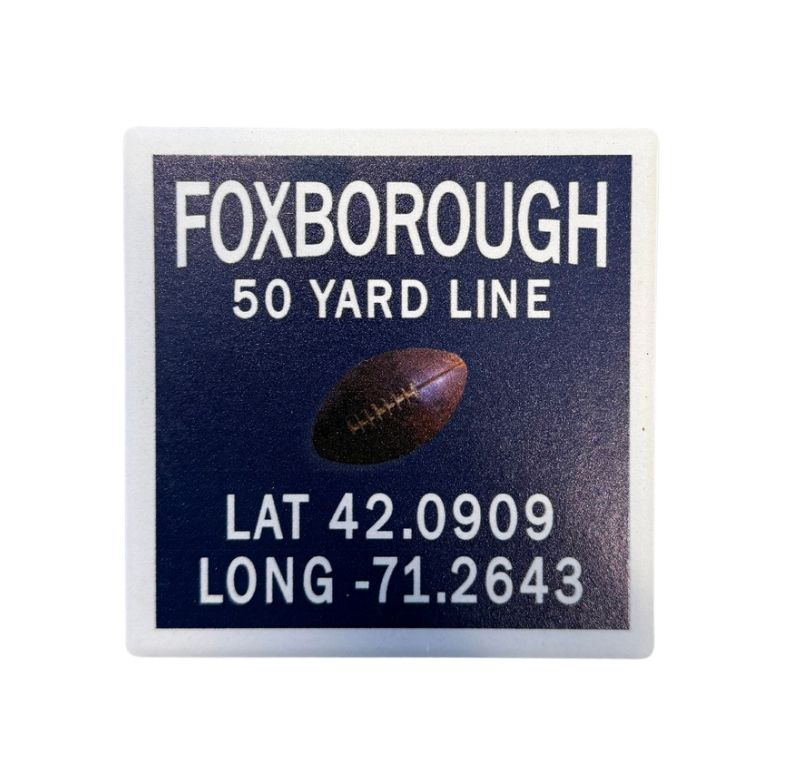 A square coaster with cork backing, predominantly in a color representing the New England Patriots, with text including 'FOXBOROUGH 50 YARD LINE' and coordinates 'LAT 42.0909 LONG -71. Available at Be Charmed