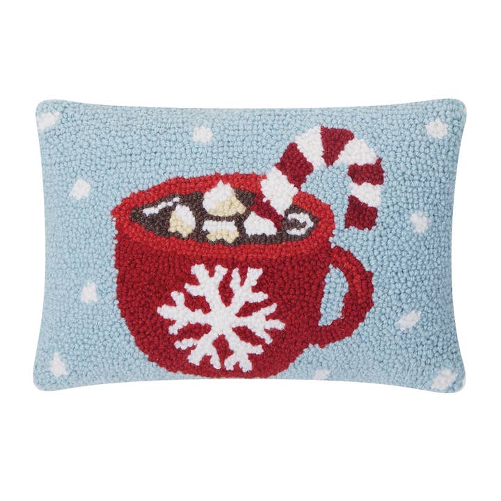 Decorative pillow with a red mug of hot chocolate, marshmallows, and a candy cane on a light blue background. Available at Be Charmed