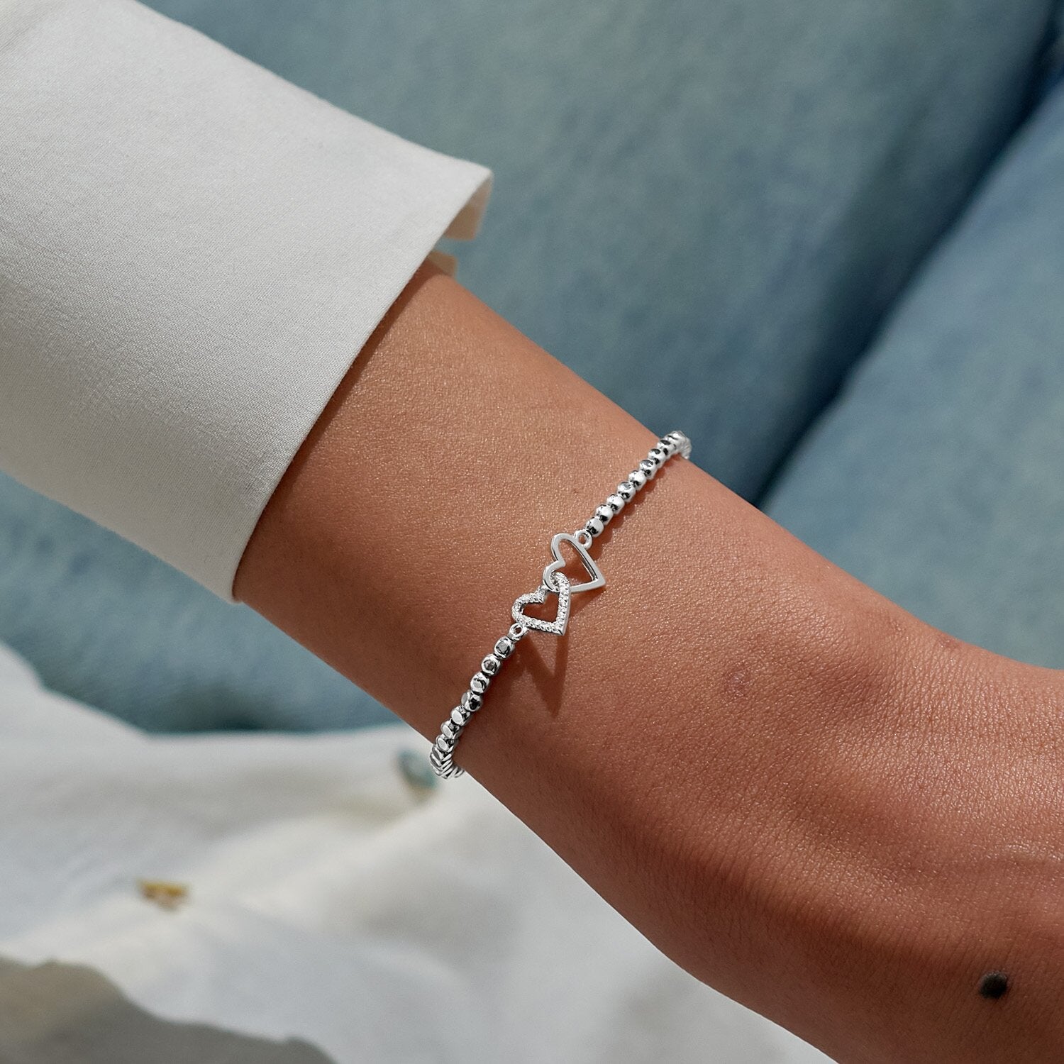 Person weraing a A Littles & Co Silver bracelet with heart design on a wrist against a blurred background. Available at Be Charmed