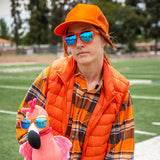 Person wearing an orange vest, plaid shirt, and sunglasses holding a pink flamingo toy on a sports field.. Available at Be Charmed