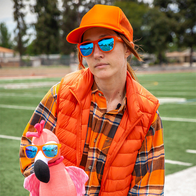 Person wearing an orange vest, plaid shirt, and sunglasses holding a pink flamingo toy on a sports field.. Available at Be Charmed