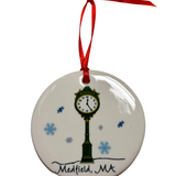Round white ornament featuring a green street clock, blue snowflakes, “Medfield, MA” lettering and a red hanging ribbon. Available at Be Charmed.

