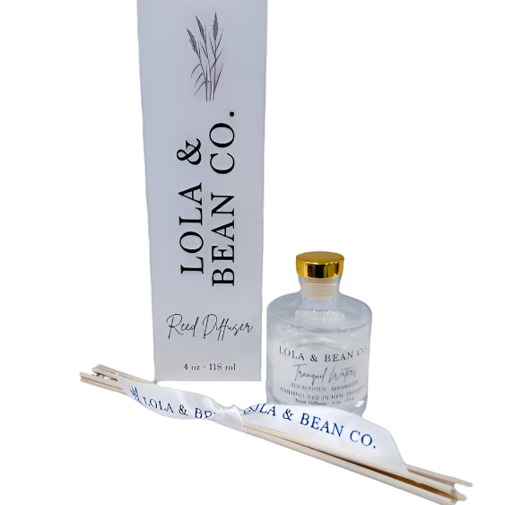 Lola & Bean 4oz Reed Diffuser – Clear glass bottle with gold collar, featuring rattan reeds and a subtle label, available at Be Charmed.